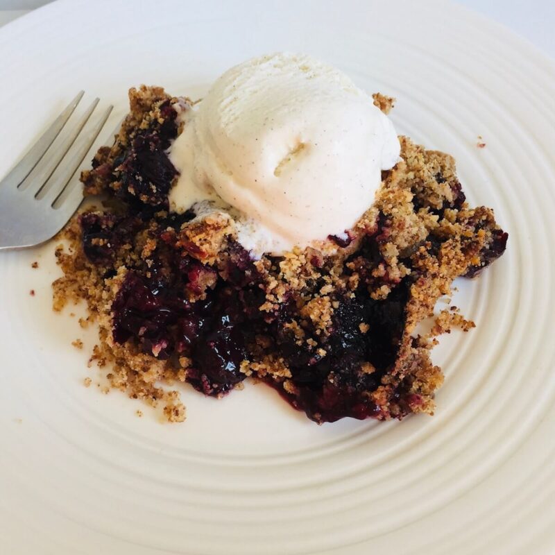 A serving of gluten-free berry crisp on a plate with vanilla ice cream melting on top next to a fork.