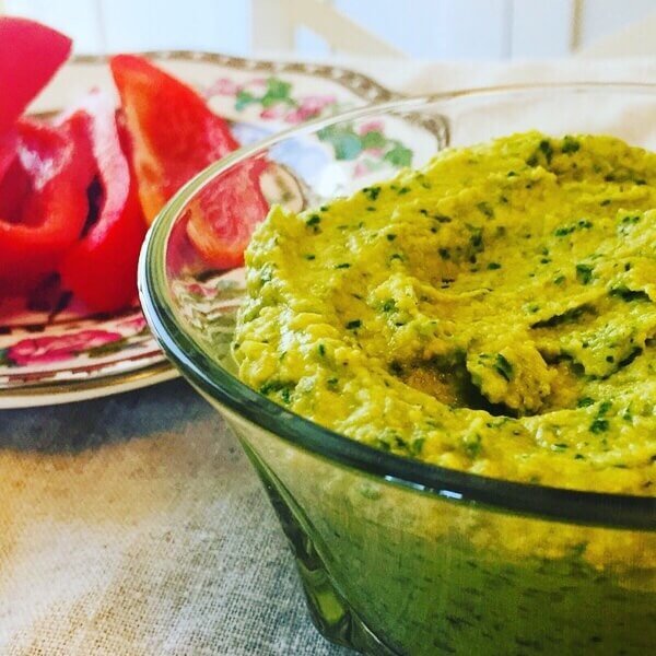 Green falafel hummus in a glass bowl next to some sliced red peppers.