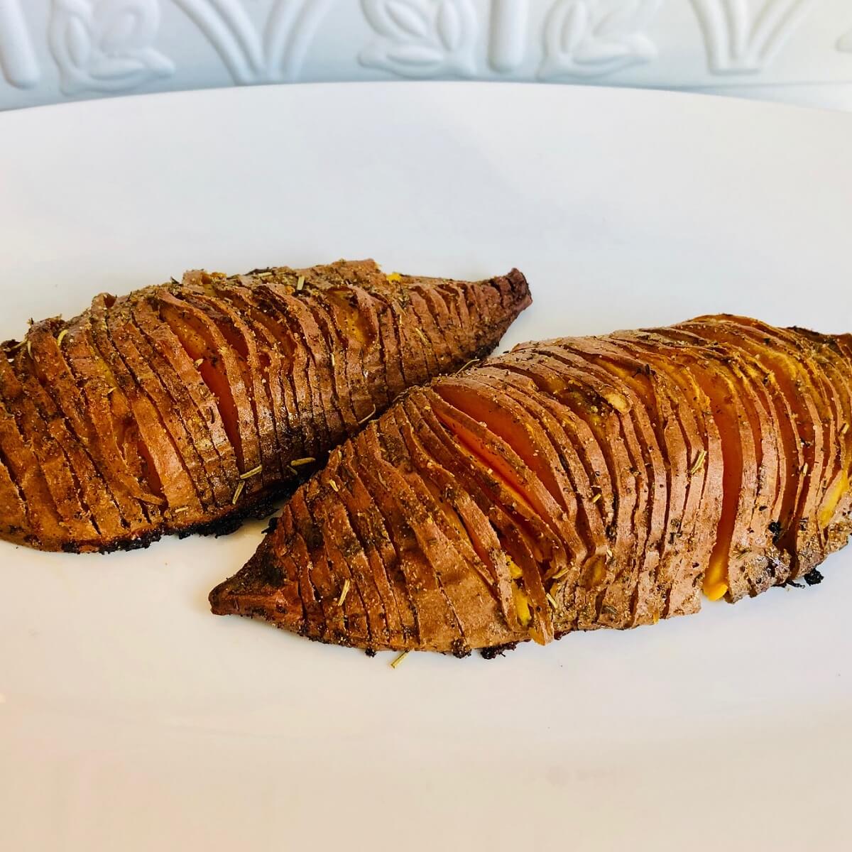 A white platter with two vegan hasselback sweet potatoes with slits cut in them.