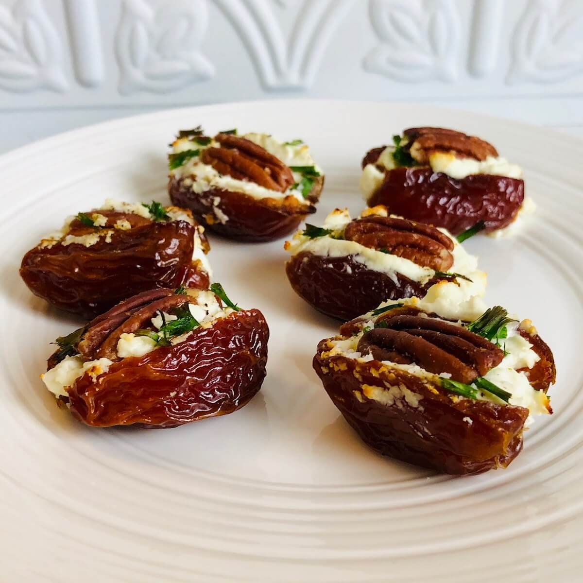 Some goat cheese and pecan stuffed dates arranged on a white platter against a white backsplash.