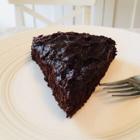 A large slice of chocolate cake on a white plate with a stainless steel fork.