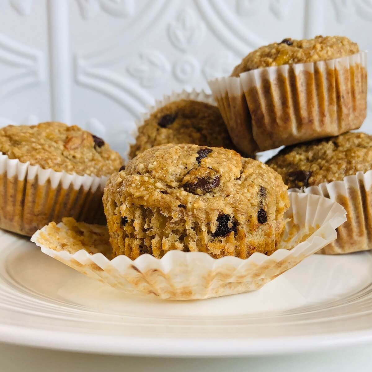 Five oatmeal banana chocolate chip muffins on a white plate against a tile back splash.