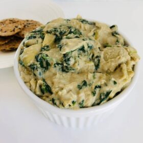 A bowl of dairy-free spinach artichoke dip next to a plate of crackers.