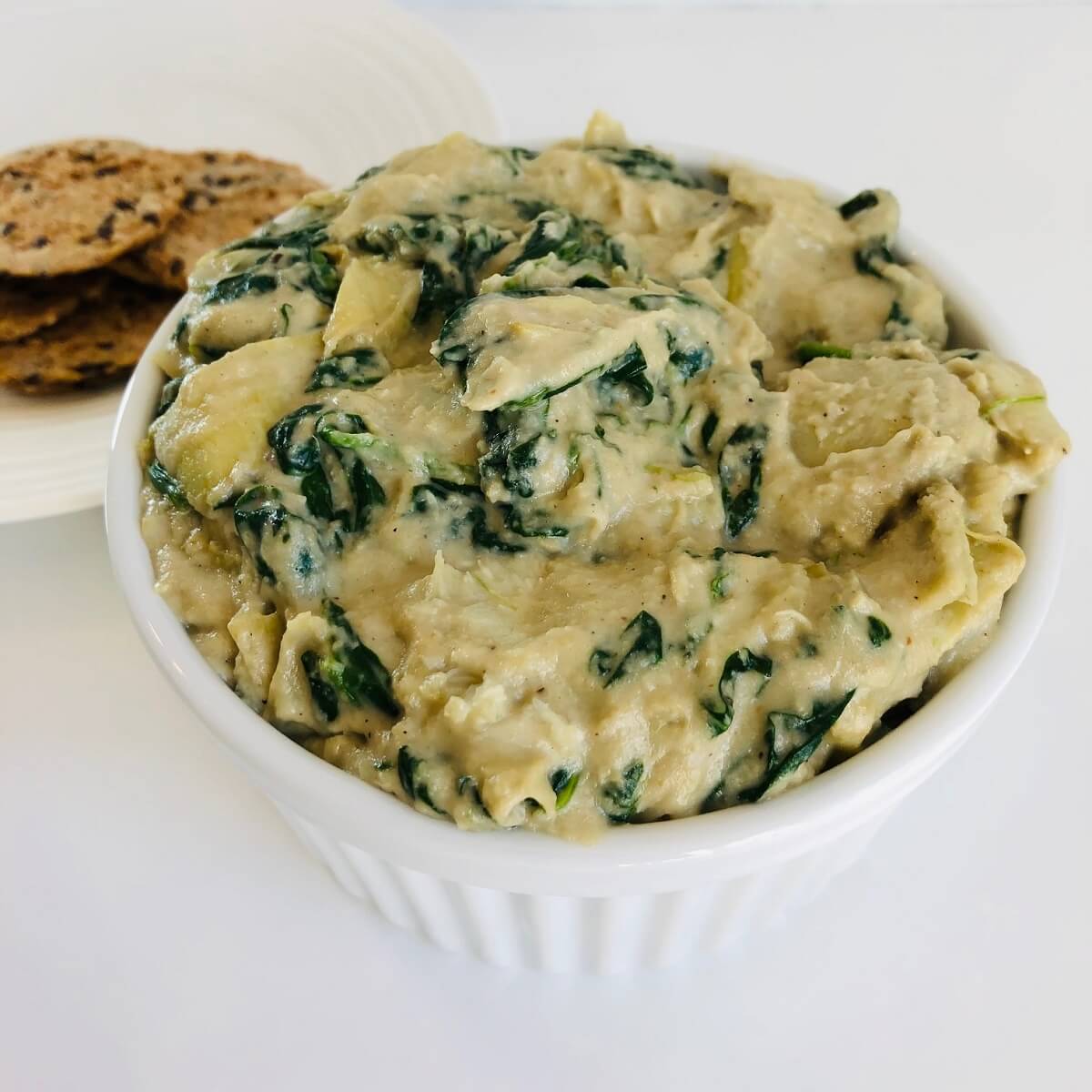 A bowl of gluten-free dairy-free spinach artichoke dip next to a plate of crackers.