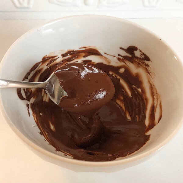 A spoon lifting some liquid chocolate mixture in a white bowl.