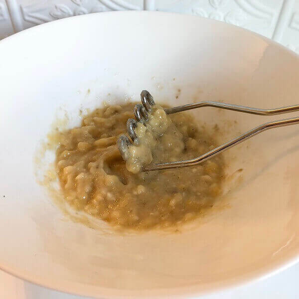 A white bowl of mashed bananas with a potato masher.