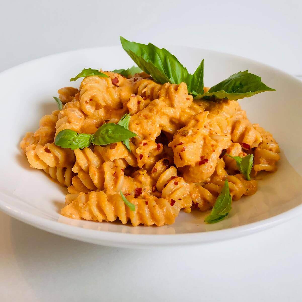 Vegan creamy tomato pasta in a white bowl garnished with basil and crushed red pepper.