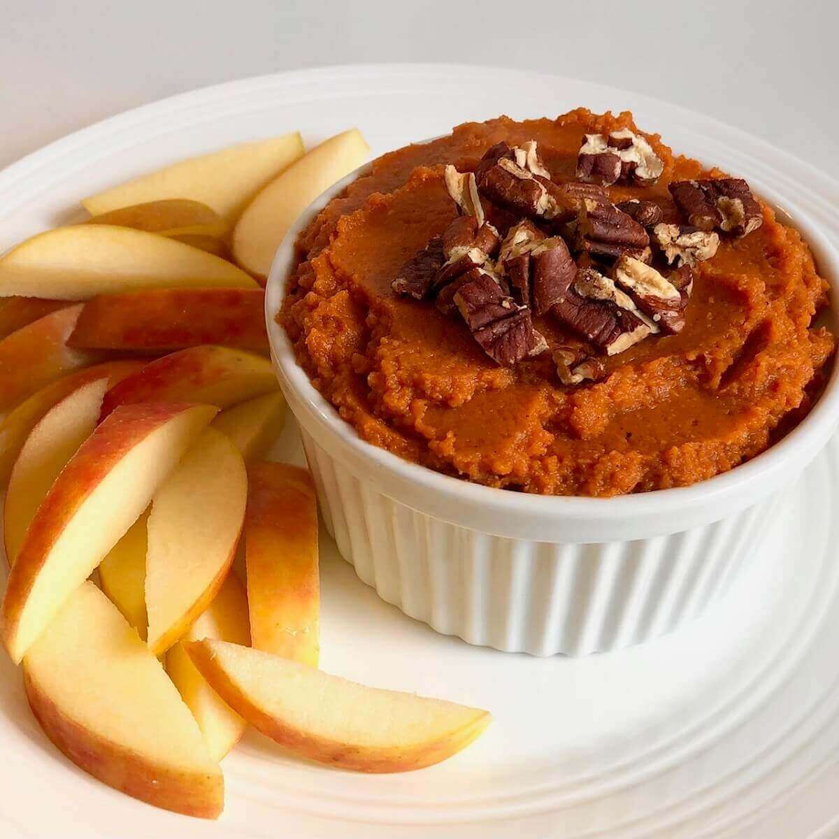 A dish filled with pumpkin spice hummus next to a pile of sliced apples.