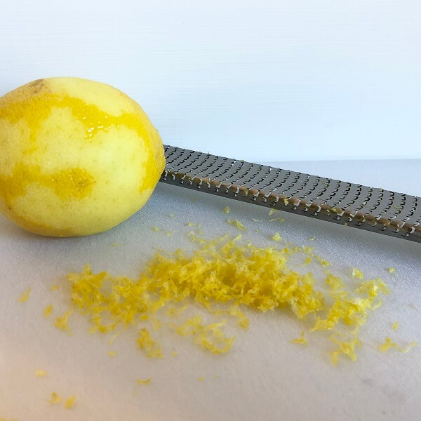Lemon zest on a cutting board next to a lemon and a metal citrus zester.