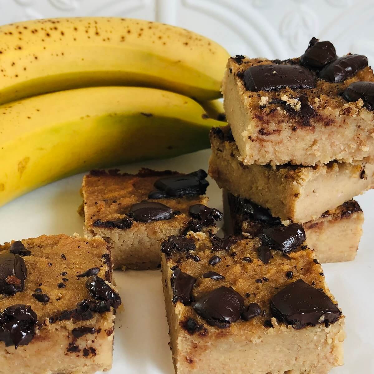 Six vegan banana blondies next to some ripe bananas.