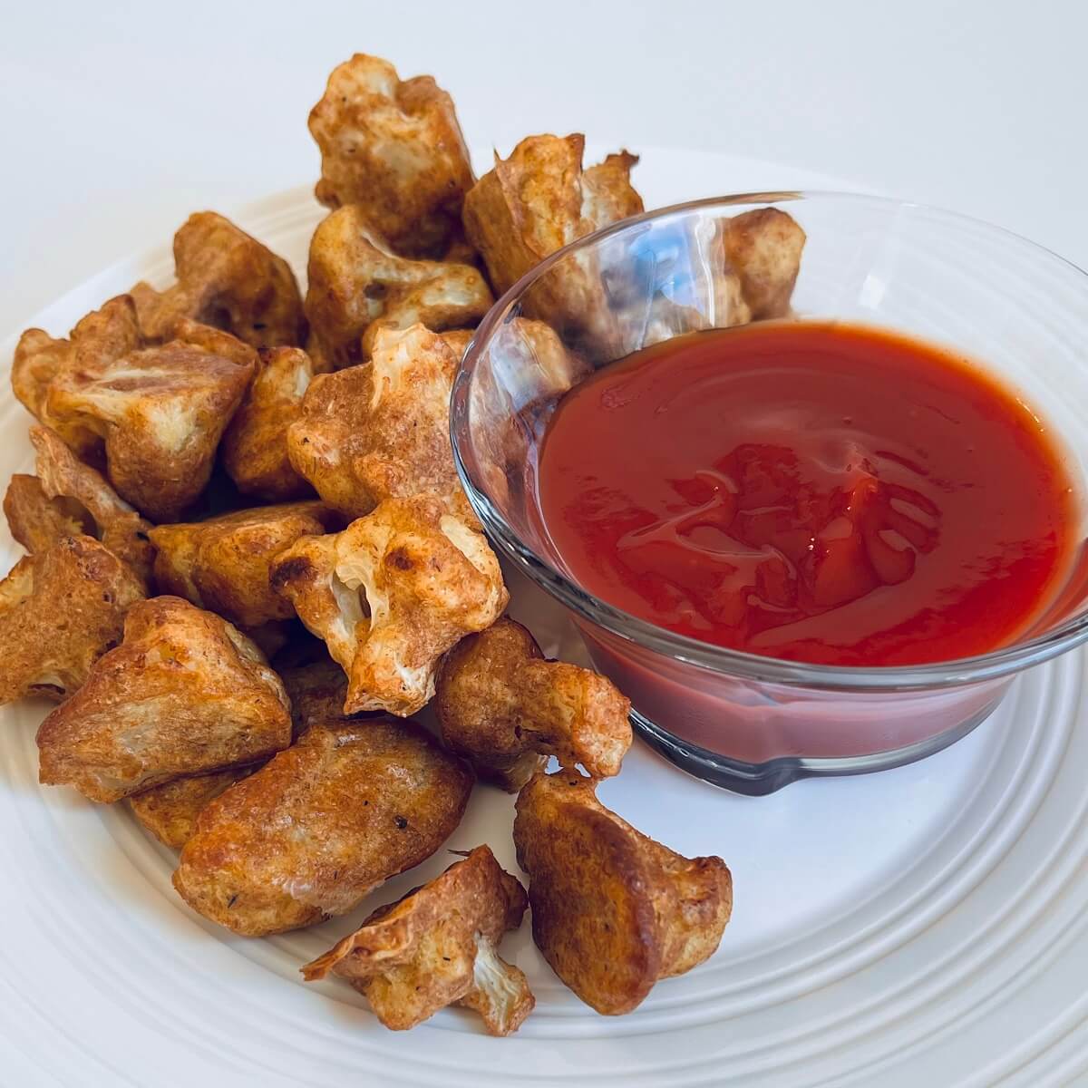 Crispy cauliflower pieces on a plate next to a glass bowl filled with ketchup.