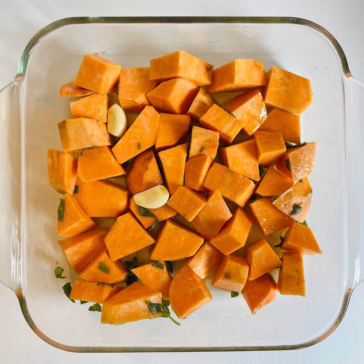 Raw sweet potato chunks, garlic cloves, and sage in a glass baking dish.