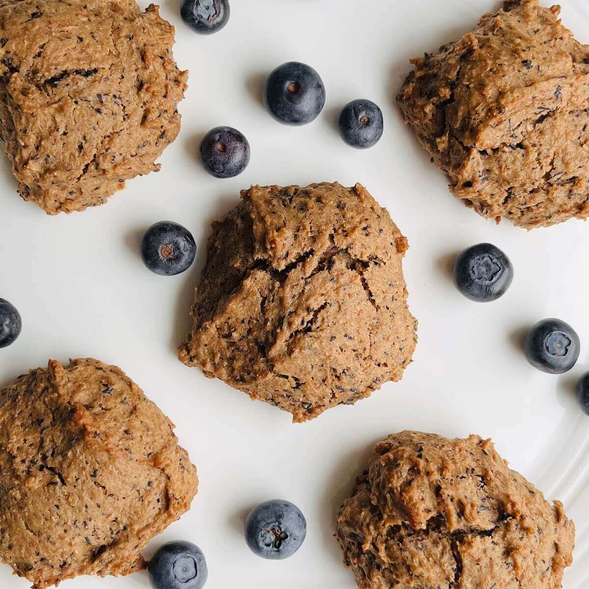 Five vegan blueberry cookies on a plate with blueberries scattered around.