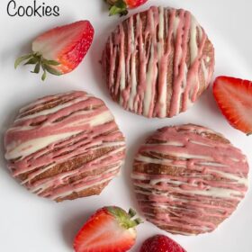 White and pink chocolate drizzled cookies on a plate next to some fresh strawberries.