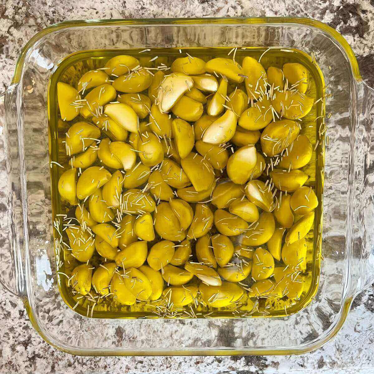 Garlic cloves, oil, and herbs in a glass baking dish.