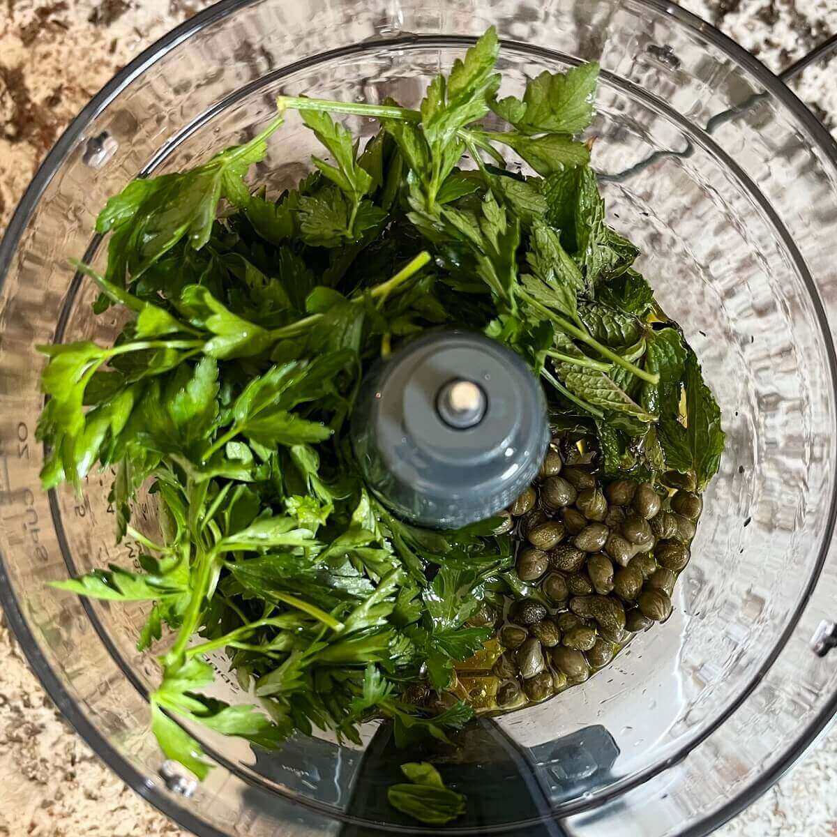 Fresh herbs and other ingredients for a sauce in a food processor.