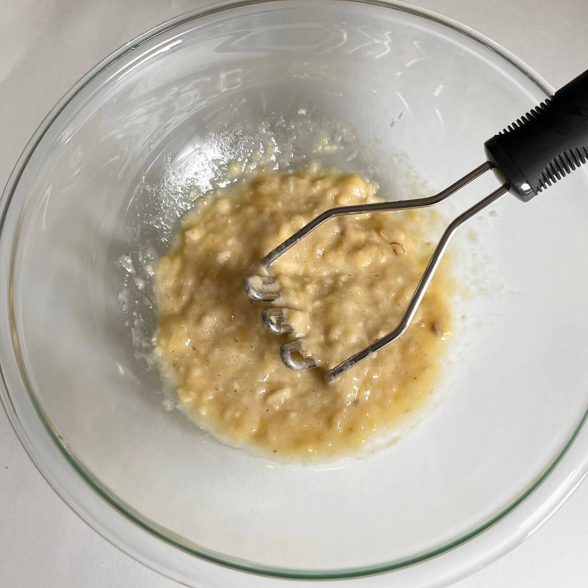 Ripe bananas being mashed by a potato masher in a glass bowl.