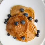 Three spelt flour pancakes with maple syrup and blueberries on a white plate.
