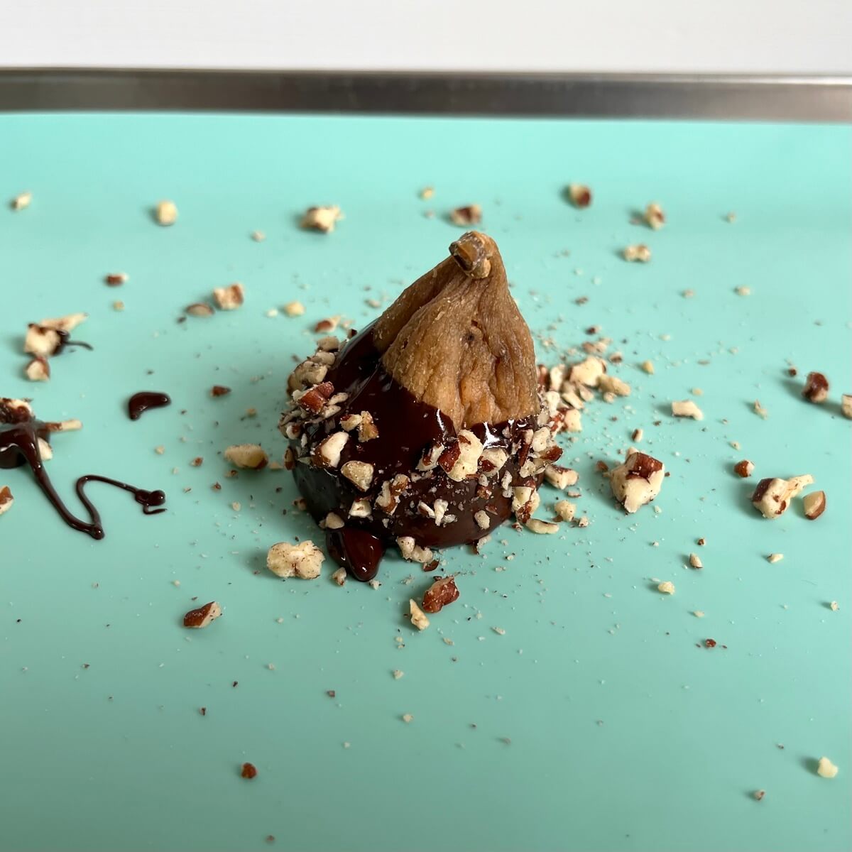 A fig dipped in chocolate and sprinkled with chopped pecans on a sheet pan lined with a silicone baking mat.