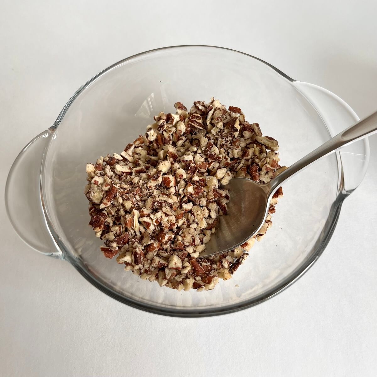 Finely chopped pecans in a glass bowl with a stainless steel spoon.