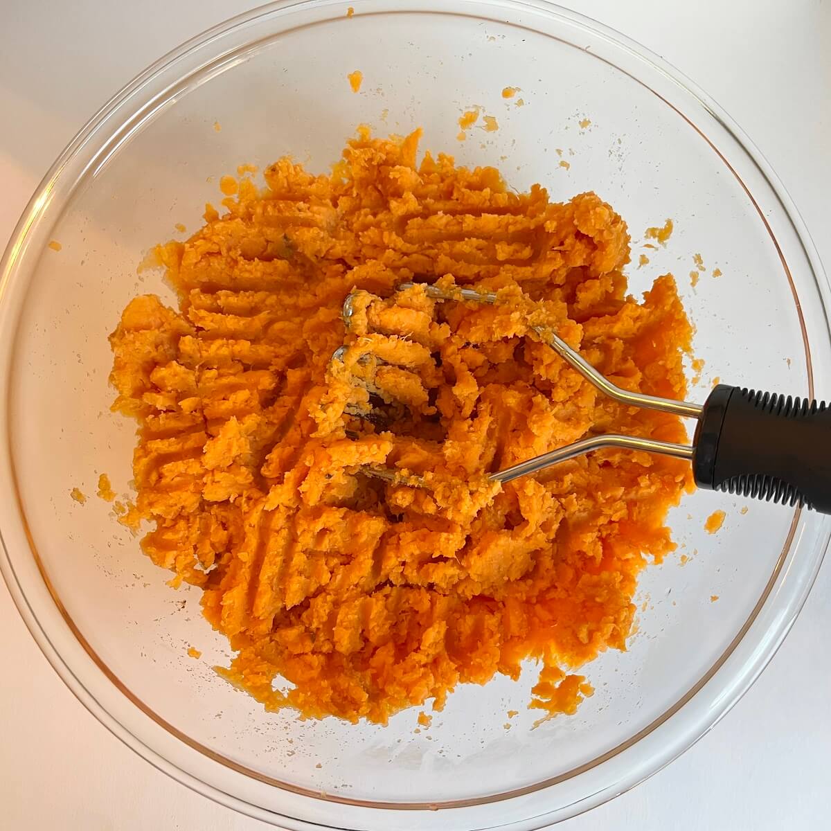 Mashed sweet potatoes in a glass bowl with a potato masher.