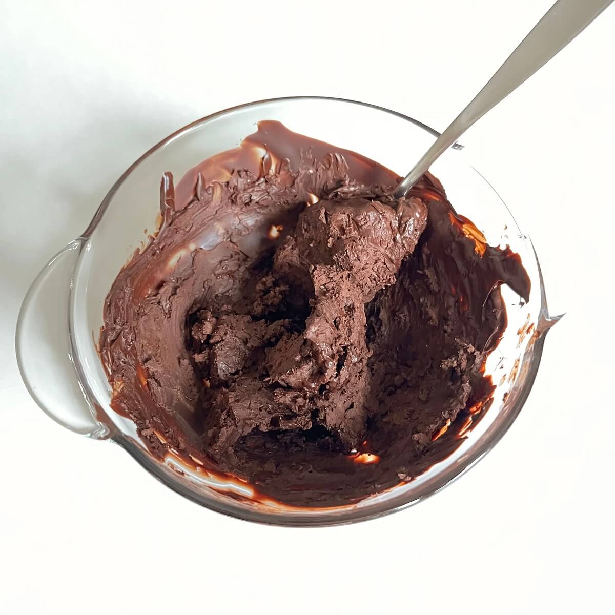A solid mixture of dark chocolate and olive oil in a glass bowl with a metal spoon.