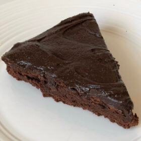 A slice of refined sugar-free chocolate cake with frosting on a white plate.