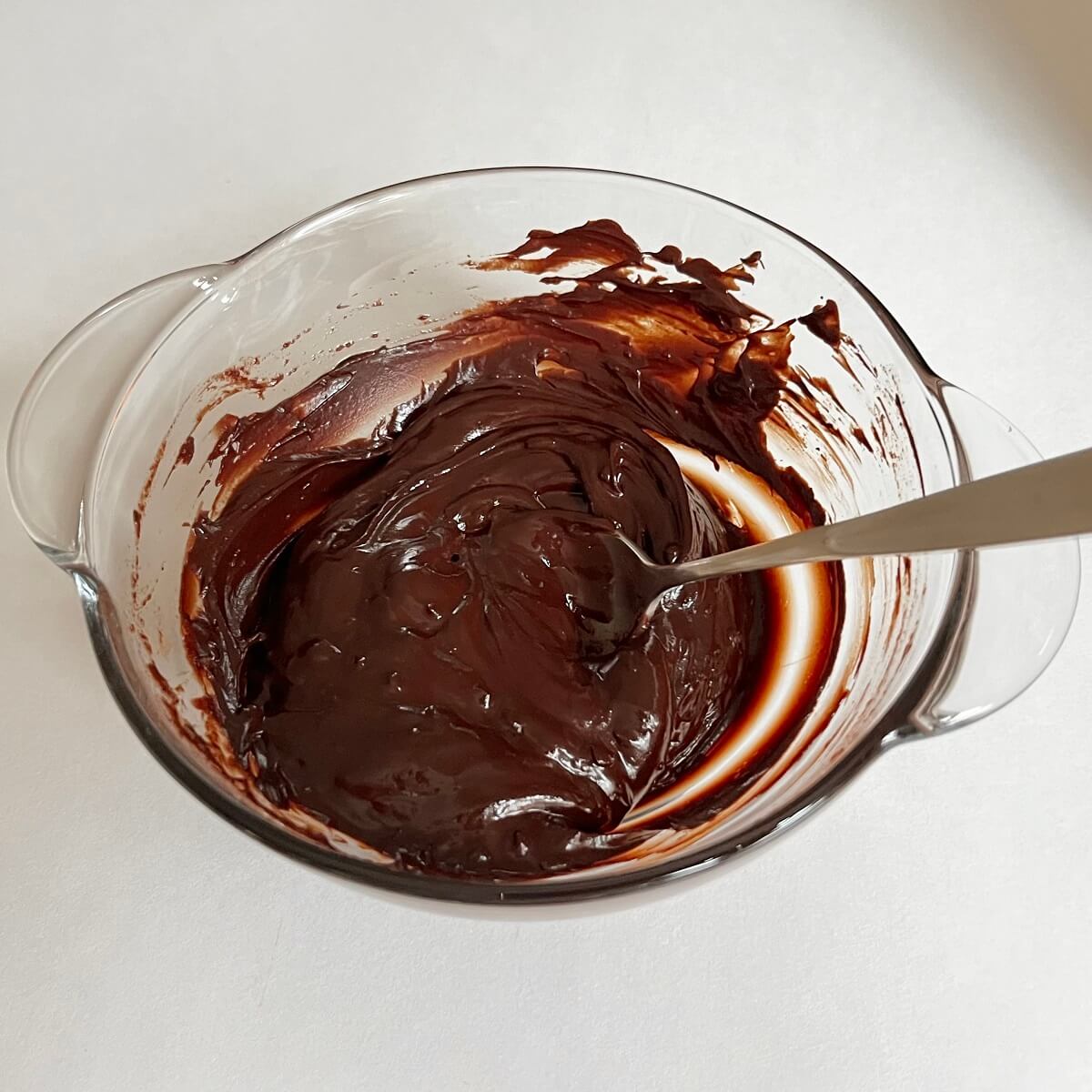 A liquid mixture of melted chocolate and lime zest and juice in a glass bowl with a metal spoon.