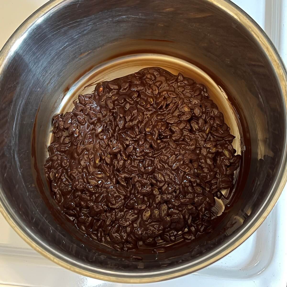 Toasted pumpkin seeds in a steel pan filled with melted chocolate.