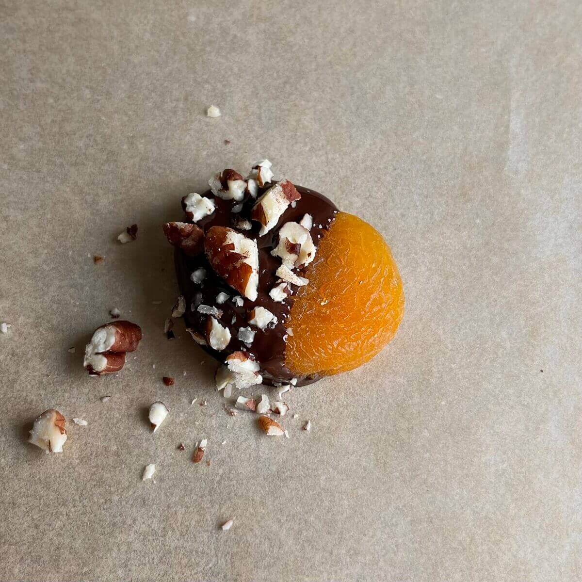 A dried apricot dipped in chocolate and sprinkled with chopped pecans.