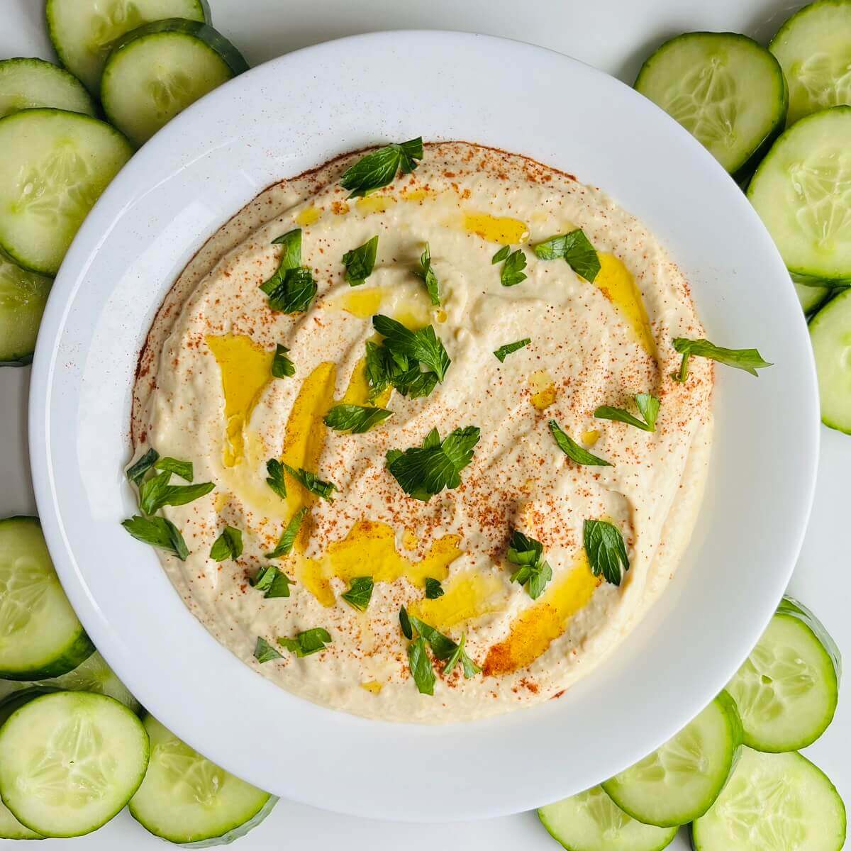 High protein hummus in a white dish surrounded by cucumber slices.