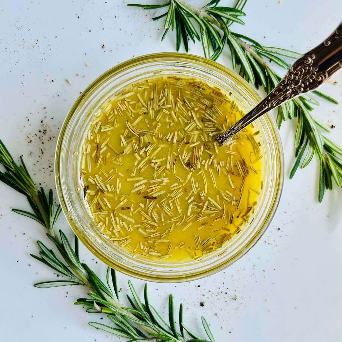 Rosemary vinaigrette in a glass bowl with an antique silver spoon surrounded by rosemary sprigs.