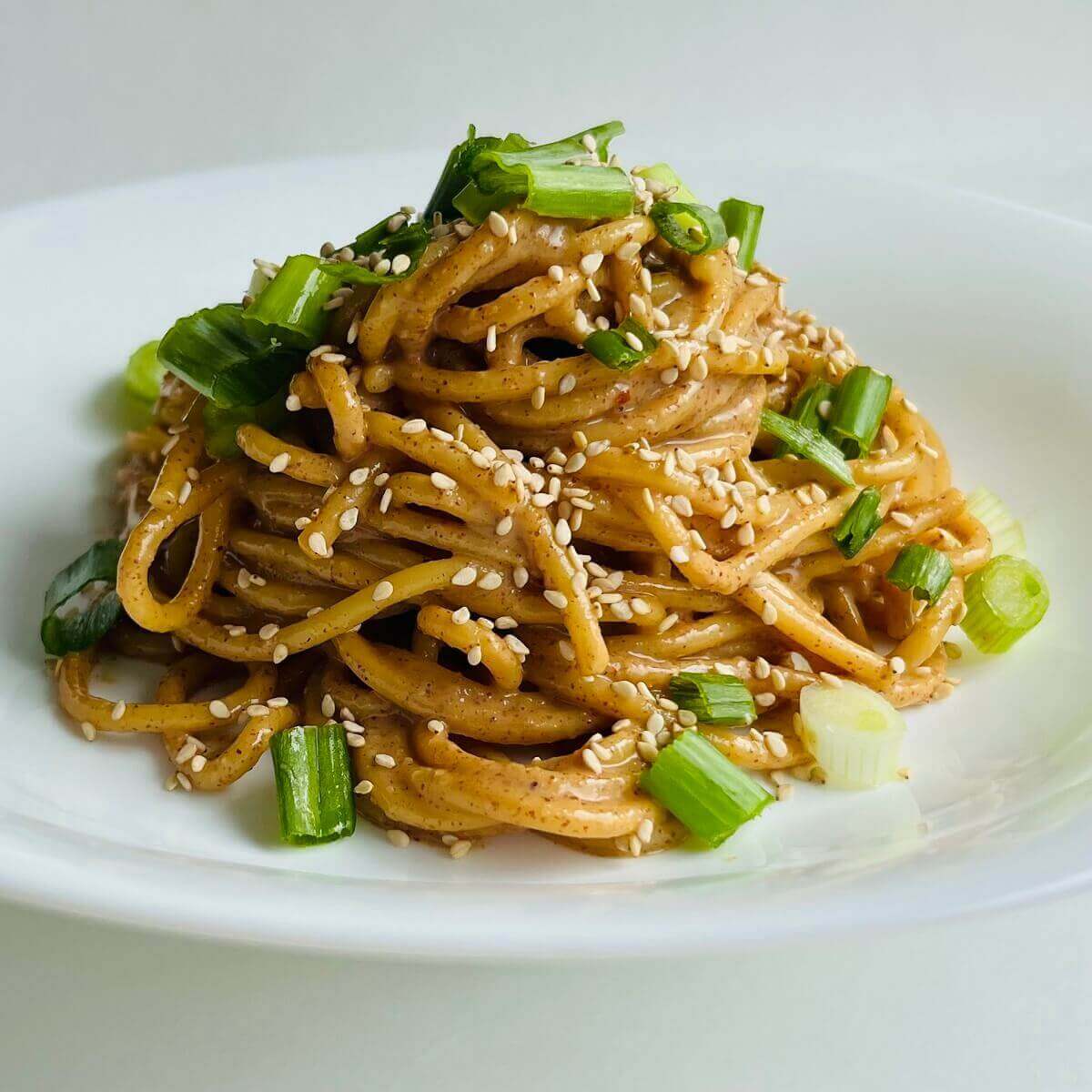 Almond butter noodles garnished with sesame seeds and chopped green onions in a white bowl.