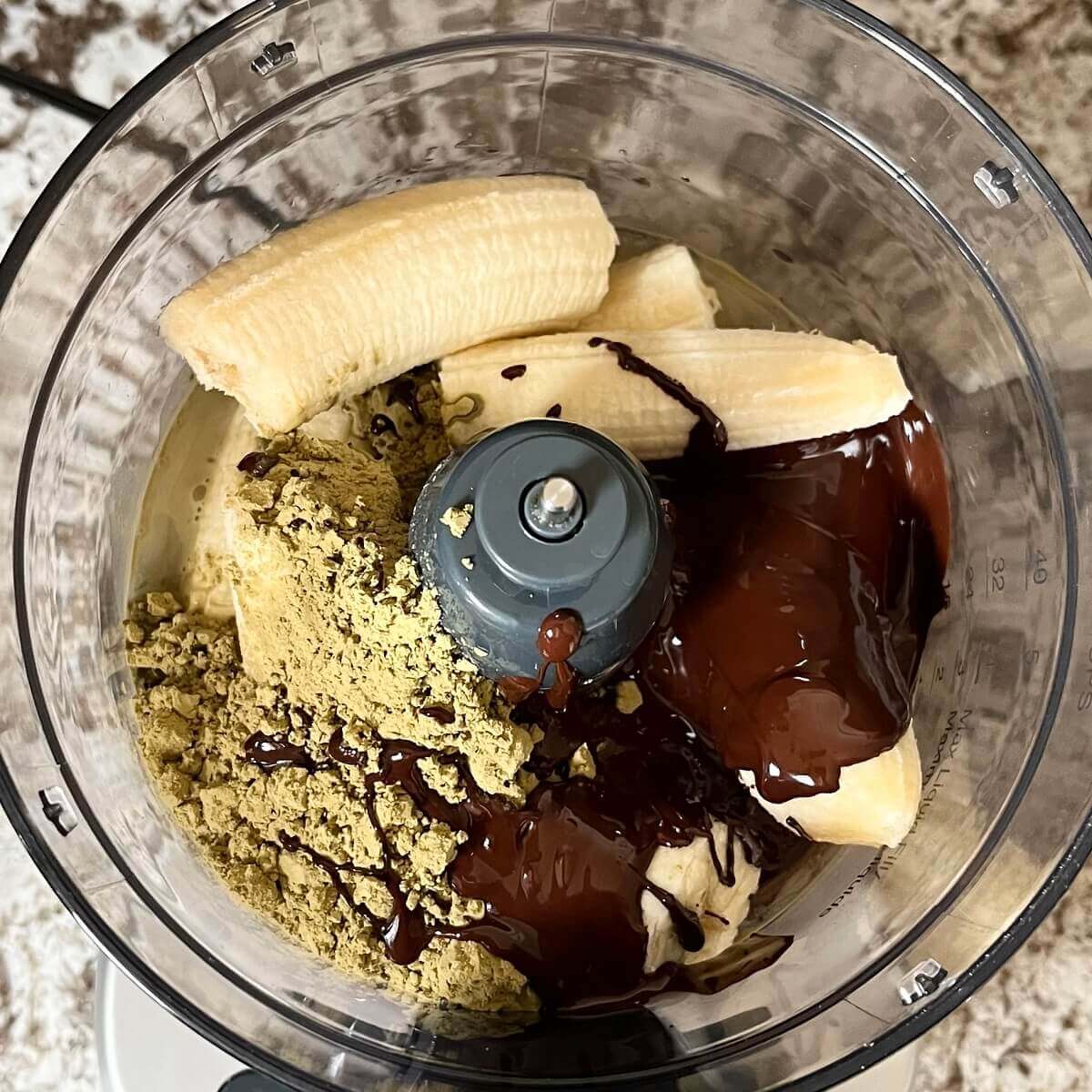Broken bananas, protein powder, melted chocolate, and other ingredients in a food processor.