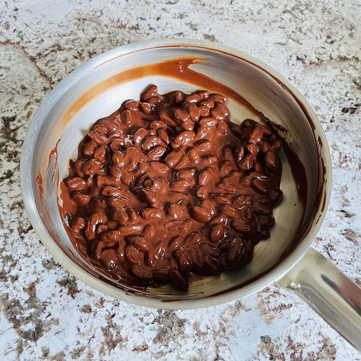 Pistachios covered in melted chocolate in a stainless steel pan.