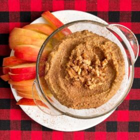 Gingerbread hummus in a glass dish next to apple slices on a white plate.