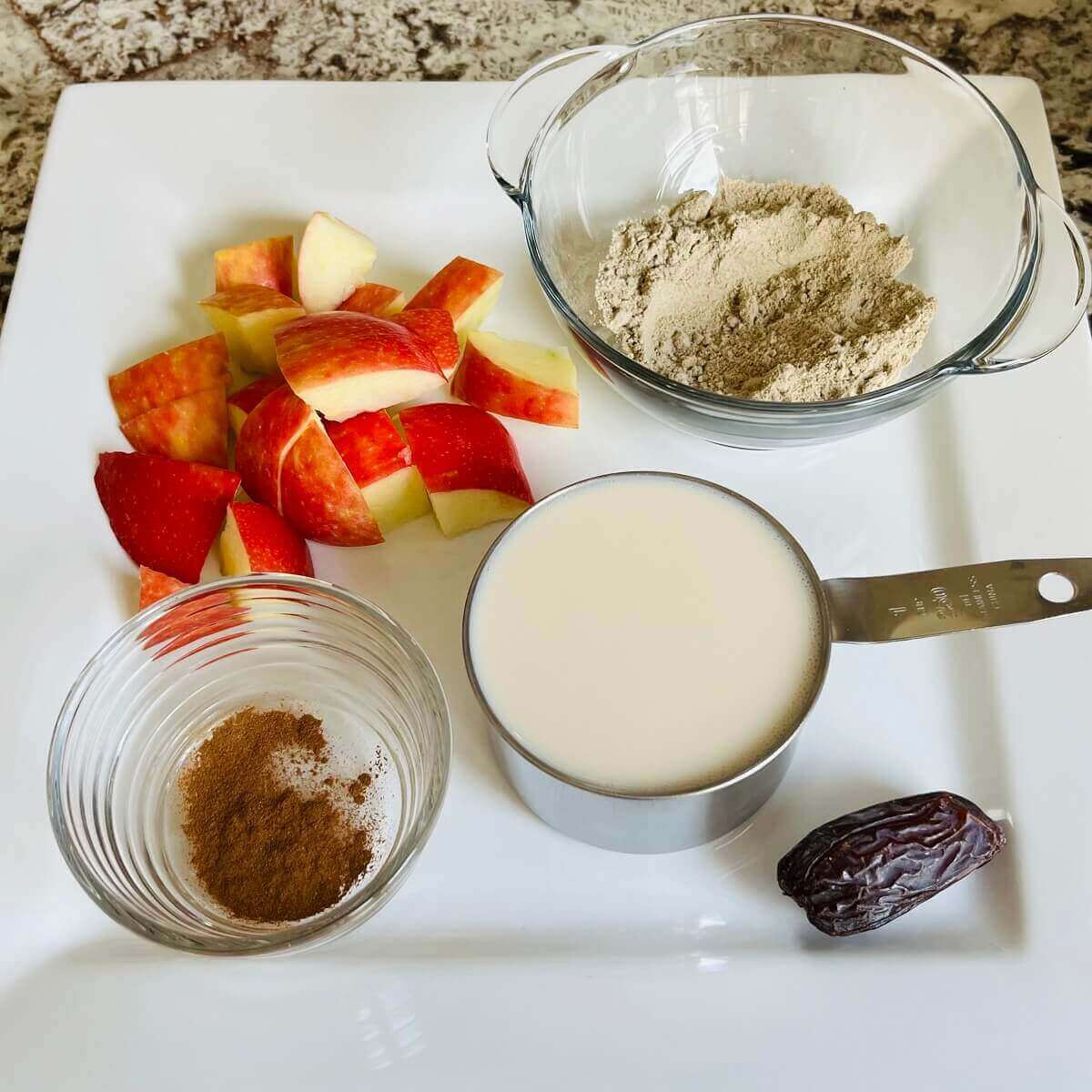 Chopped apples, a bowl of protein powder, cinnamon in a bowl, a date, and a measuring cup of soy milk on a white plate.