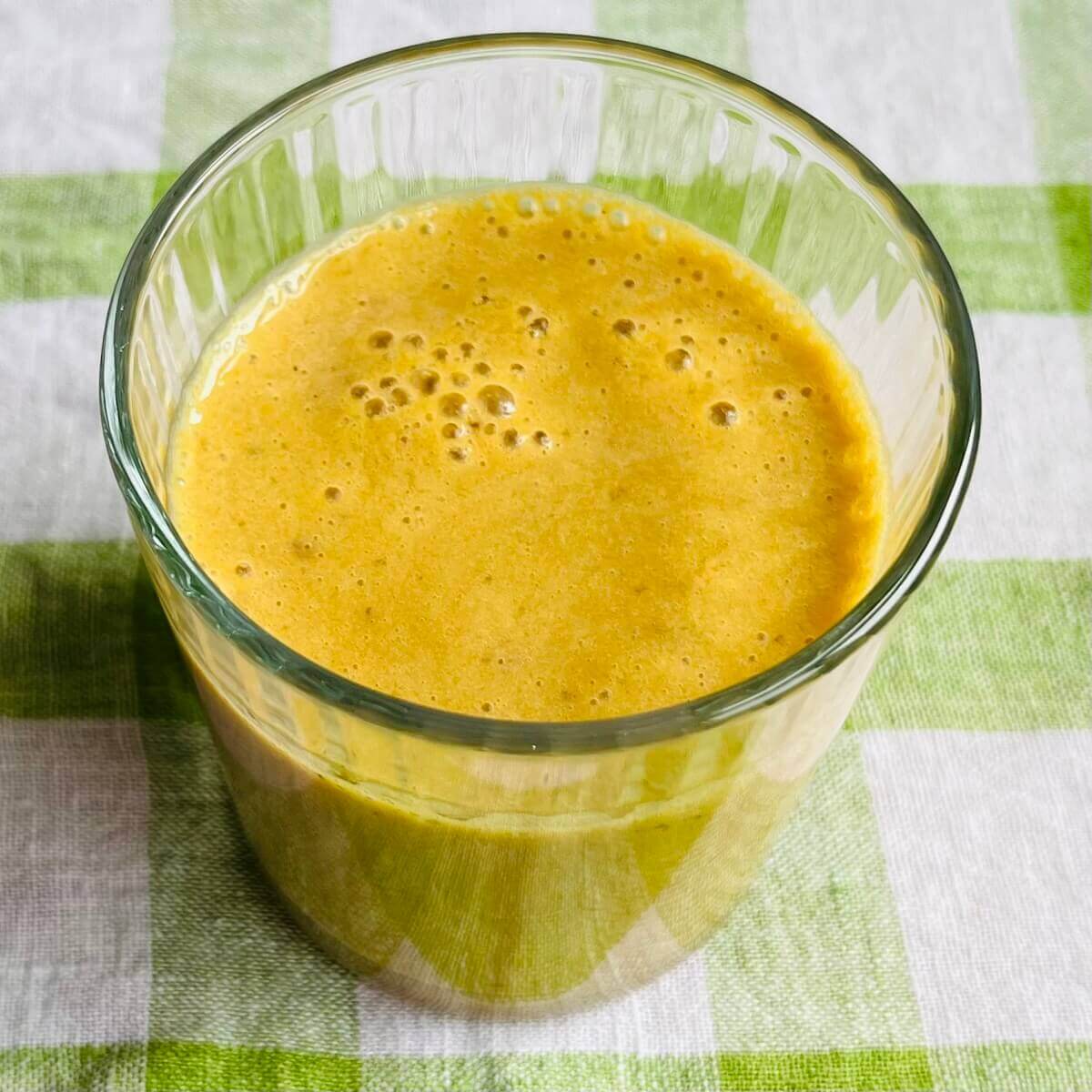 A banana turmeric smoothie in a glass on a green and white gingham tea towel.