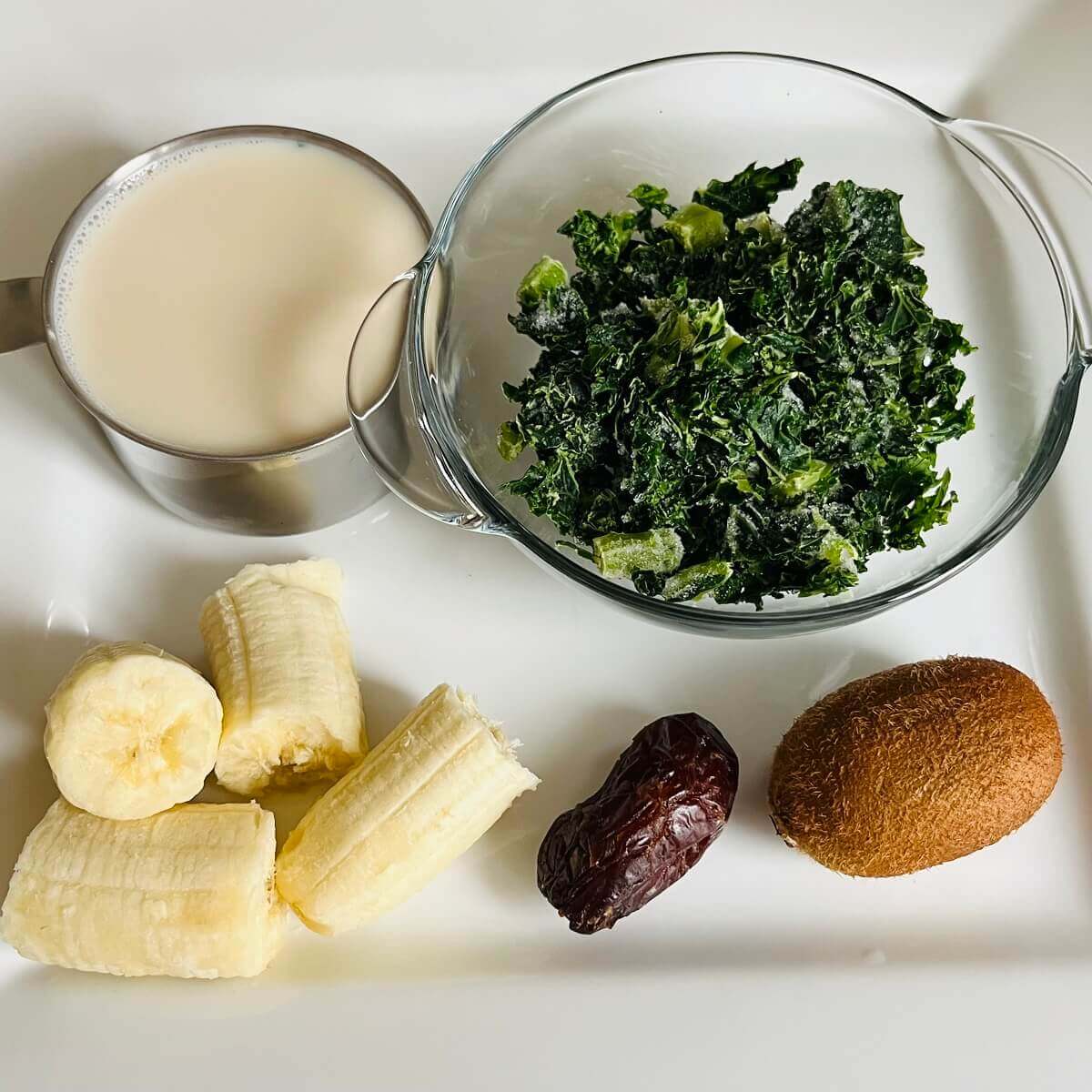 Chopped kale in a bowl, a cup of milk, banana pieces, a date, and a kiwi on a white plate.