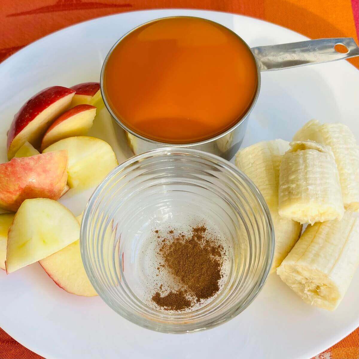 A cup of carrot juice, apple chunks, banana pieces, and a bowl of cinnamon on a white plate.