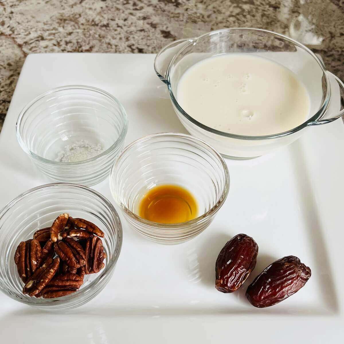 Pecans, dates, and bowls of vanilla extract, sea salt, and almond milk on a square plate.