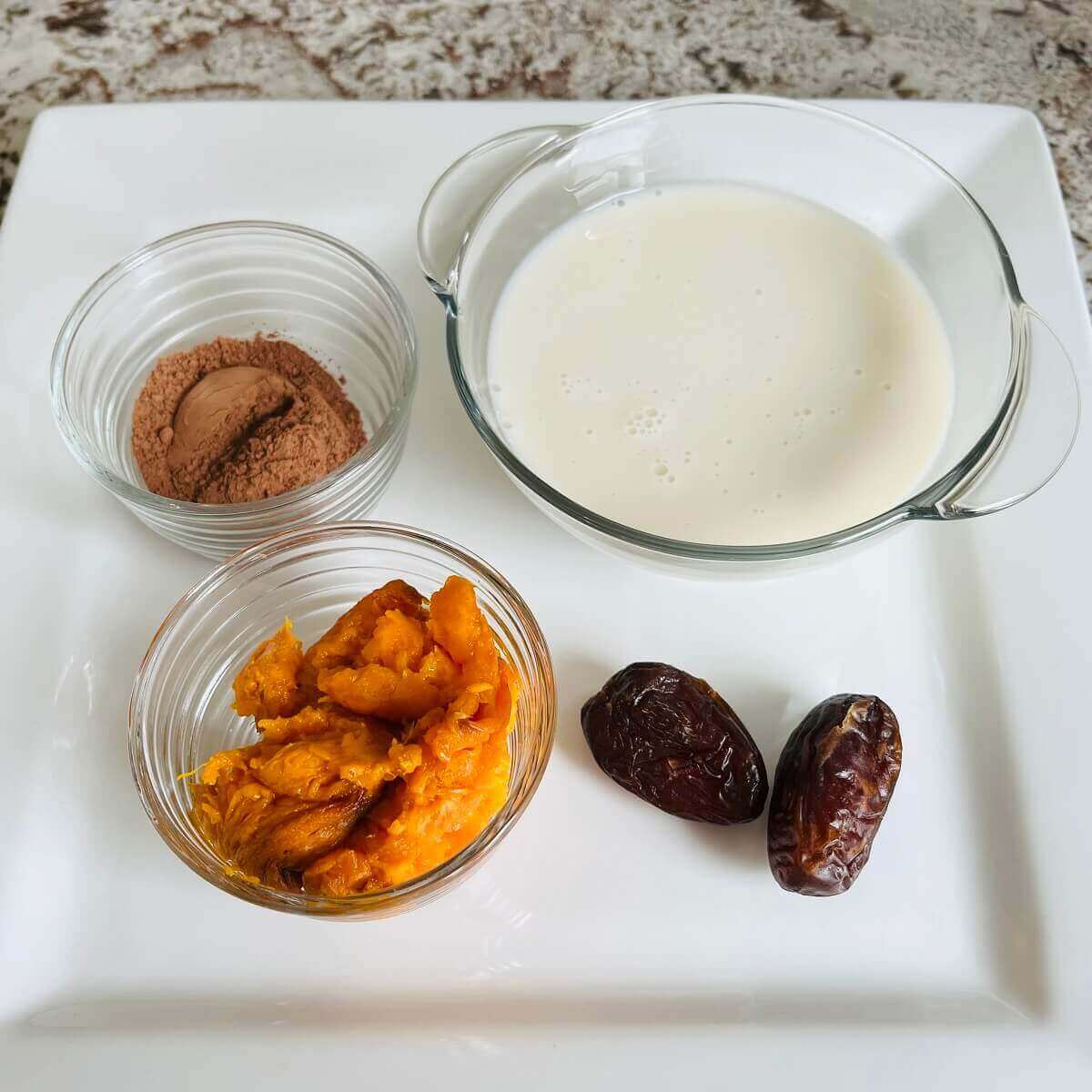Two dates and bowls of sweet potato, cocoa powder, and almond milk on a white square plate.