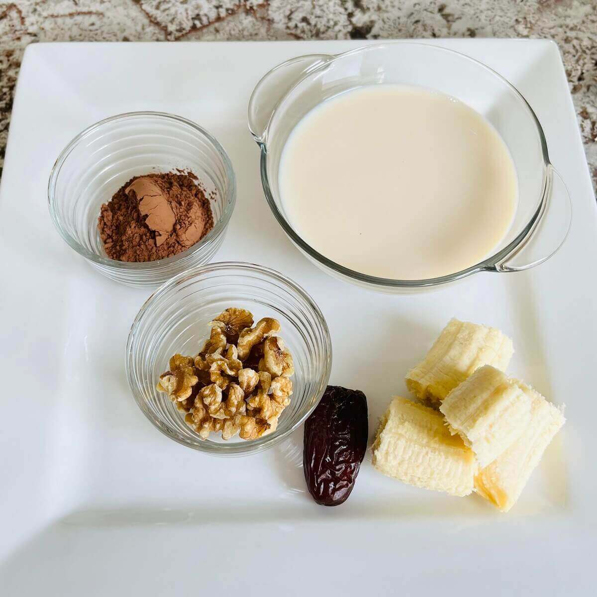 Walnuts, banana pieces, a date, and bowls of soy milk and cocoa powder on a plate.