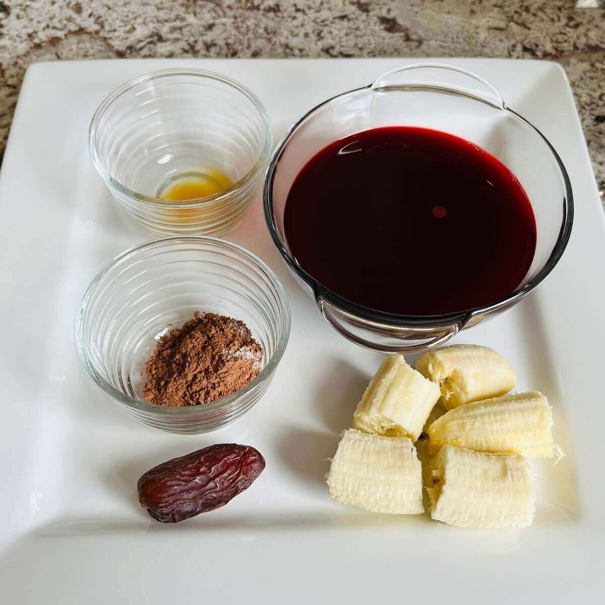 Banana chunks, a date, and bowls of beet juice, vanilla extract, and cocoa powder on a platter.