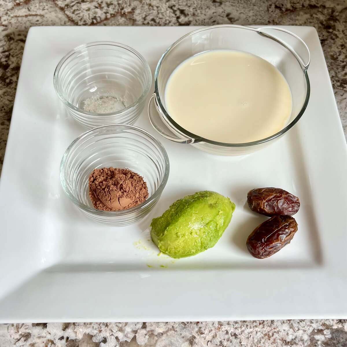 Dates, avocado, and bowls of soy milk, cocoa powder, and sea salt on a platter.
