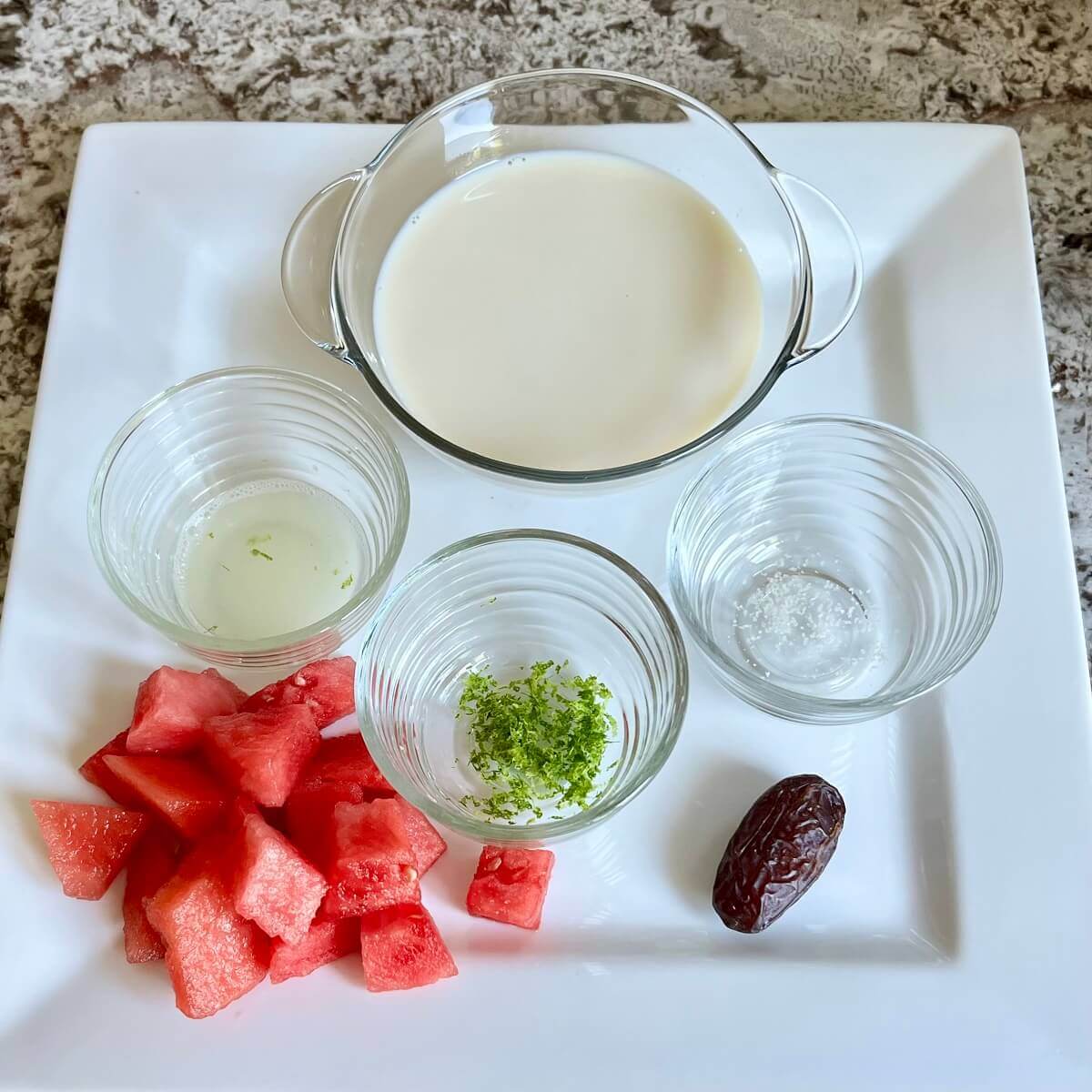 Chopped watermelon, a date, and bowls of lime zest, lime juice, sea salt, and soy milk on a platter.