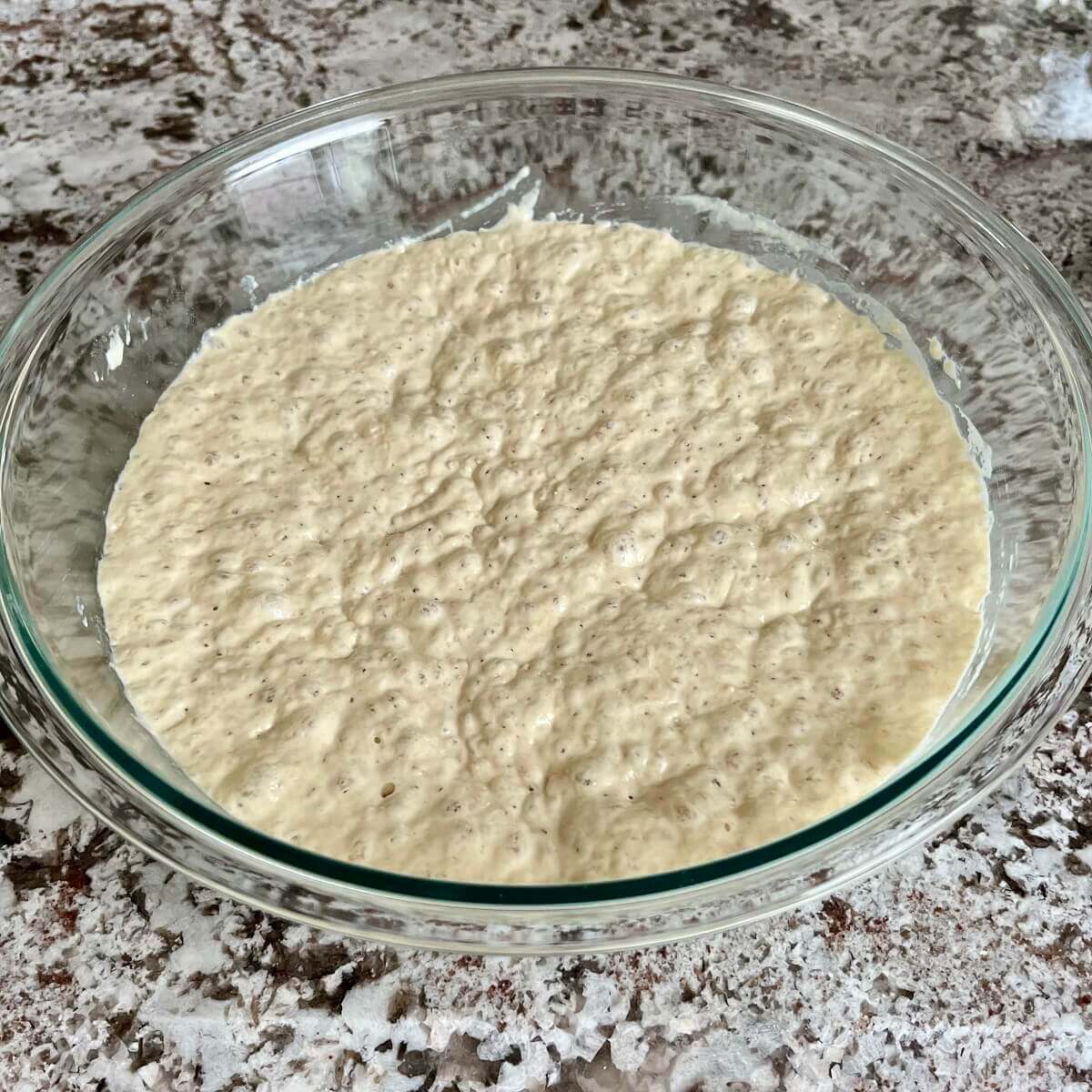 Fully risen no-knead bread dough in a glass bowl.