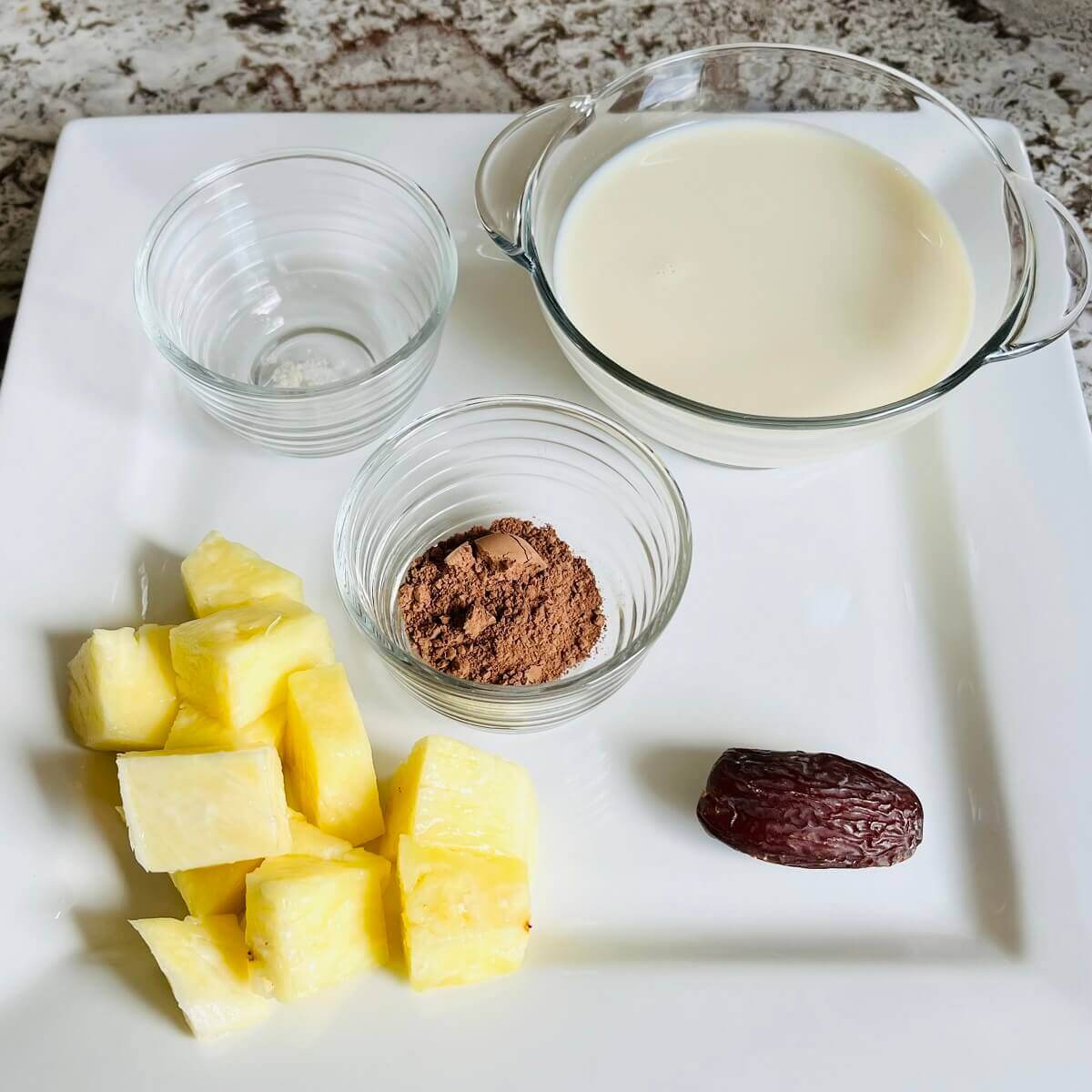 Chopped pineapple, a date, and bowls of soy milk, cocoa powder, and sea salt on a platter.