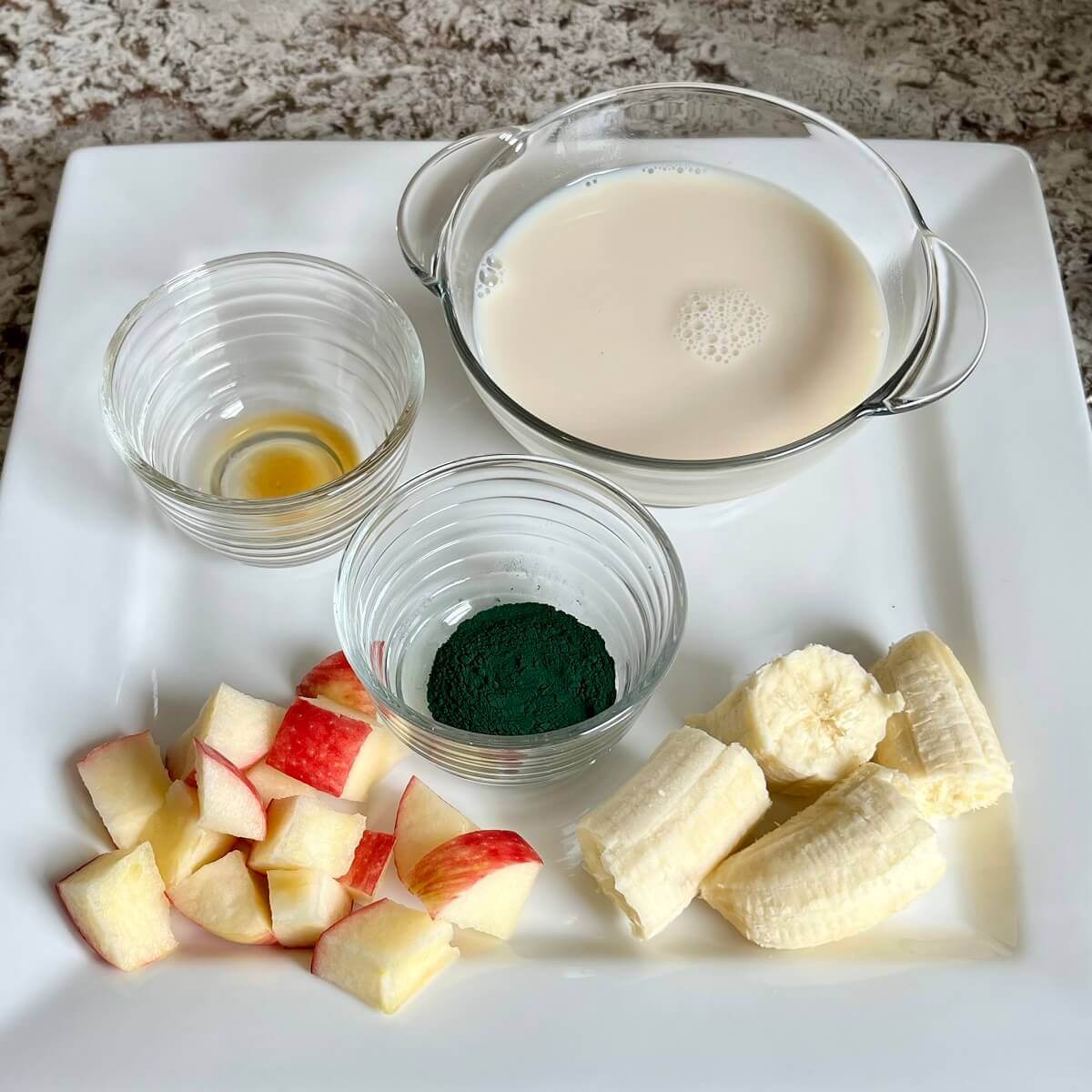Chopped apple, banana, and bowls of spirulina powder, vanilla, and soy milk on a platter.