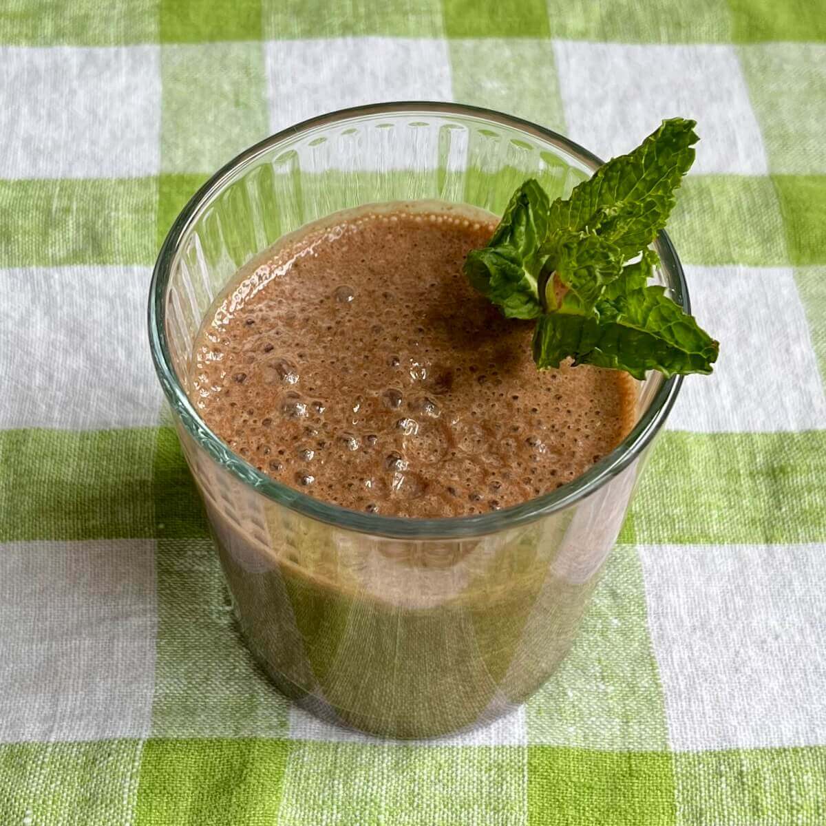 A cocoa mint smoothie in a glass on a green gingham cloth.